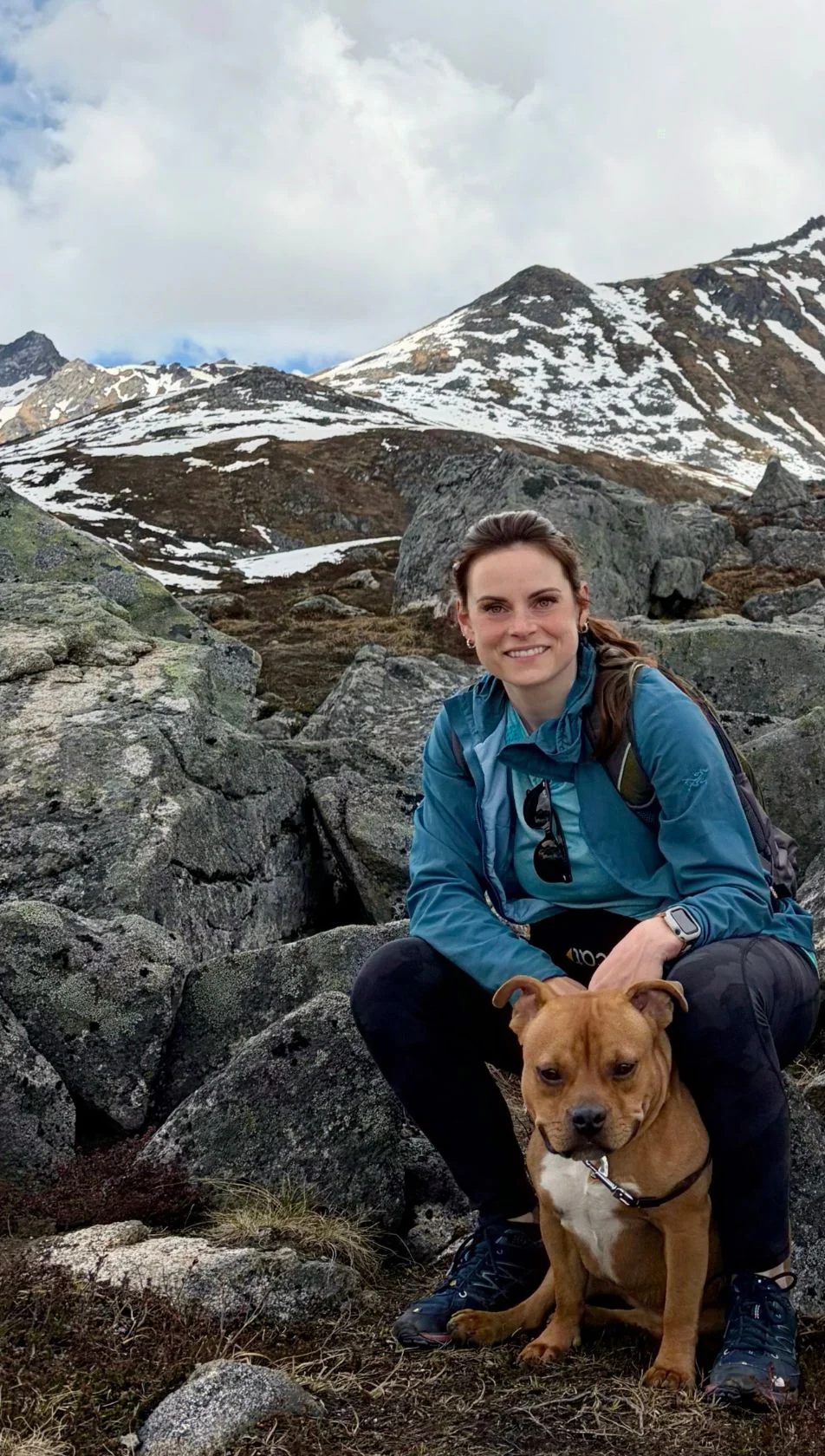 Casta Townsley crouching among mossy boulders with her dog, snow-dusted mountains behind her.
