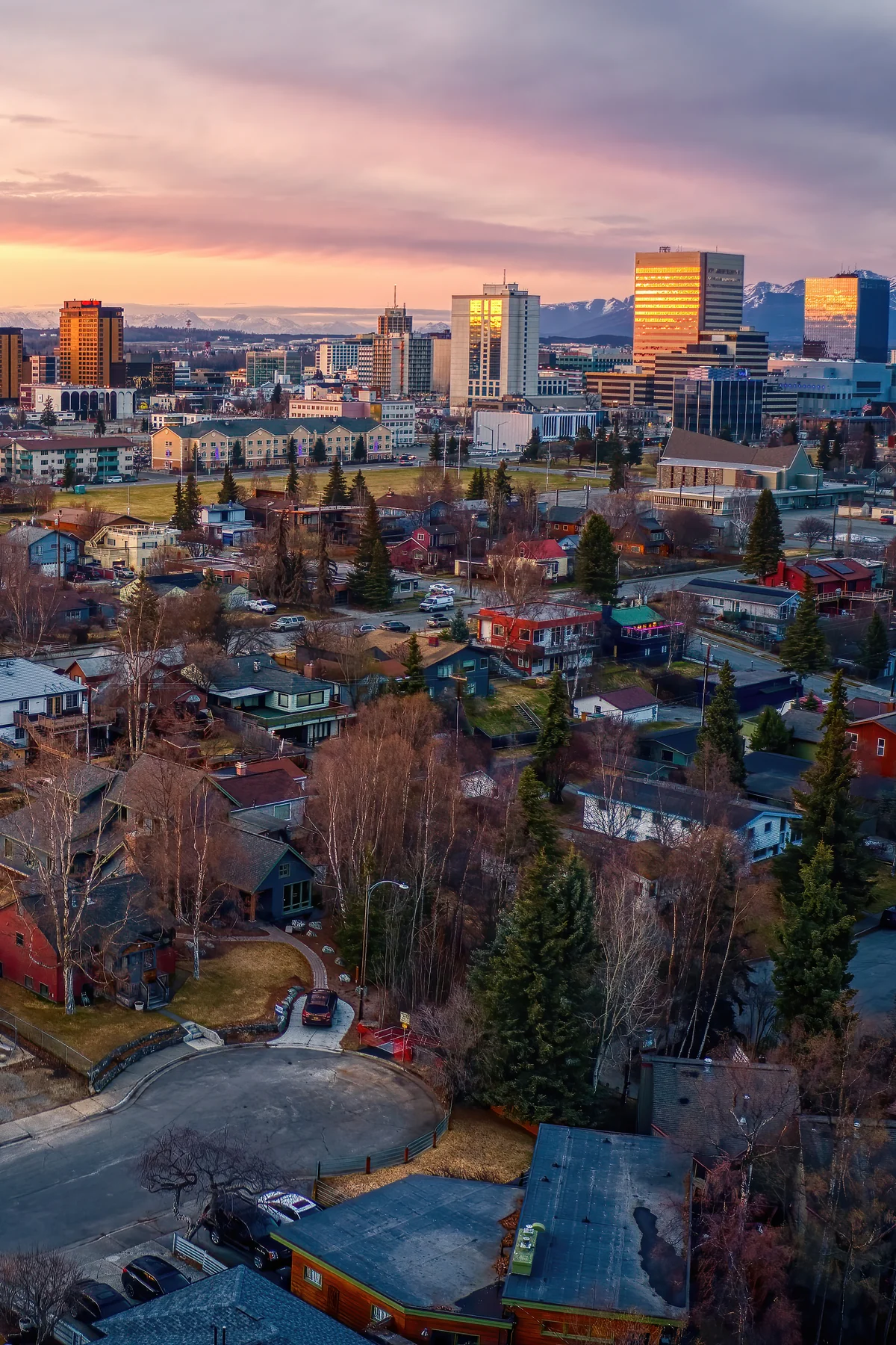 Downtown Anchorage at sunset from above, the high-rises and neighborhoods glowing beneath the snow-capped Chugach Mountains.