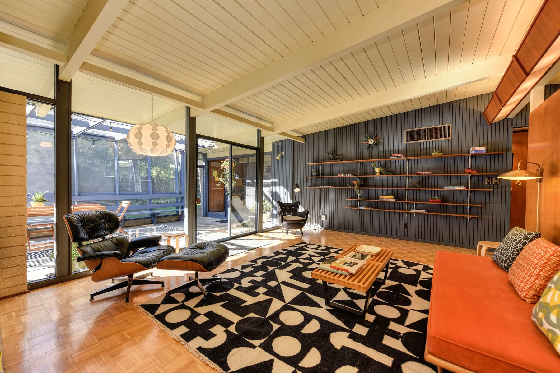 A midcentury modern room with a beamed wood ceiling, low seating and a wall of windows.